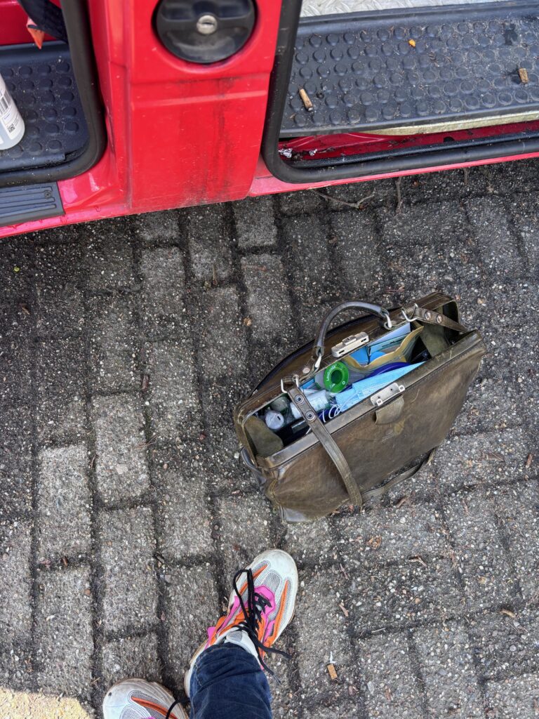 Van bovenaf gezien: een paar sneakers, een geopende dokterstas met verbandmateriaal en de rode zijkant van een busje.
