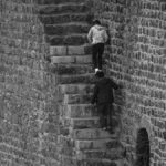 Two individuals climbing steep, narrow staircase on historical stone wall.