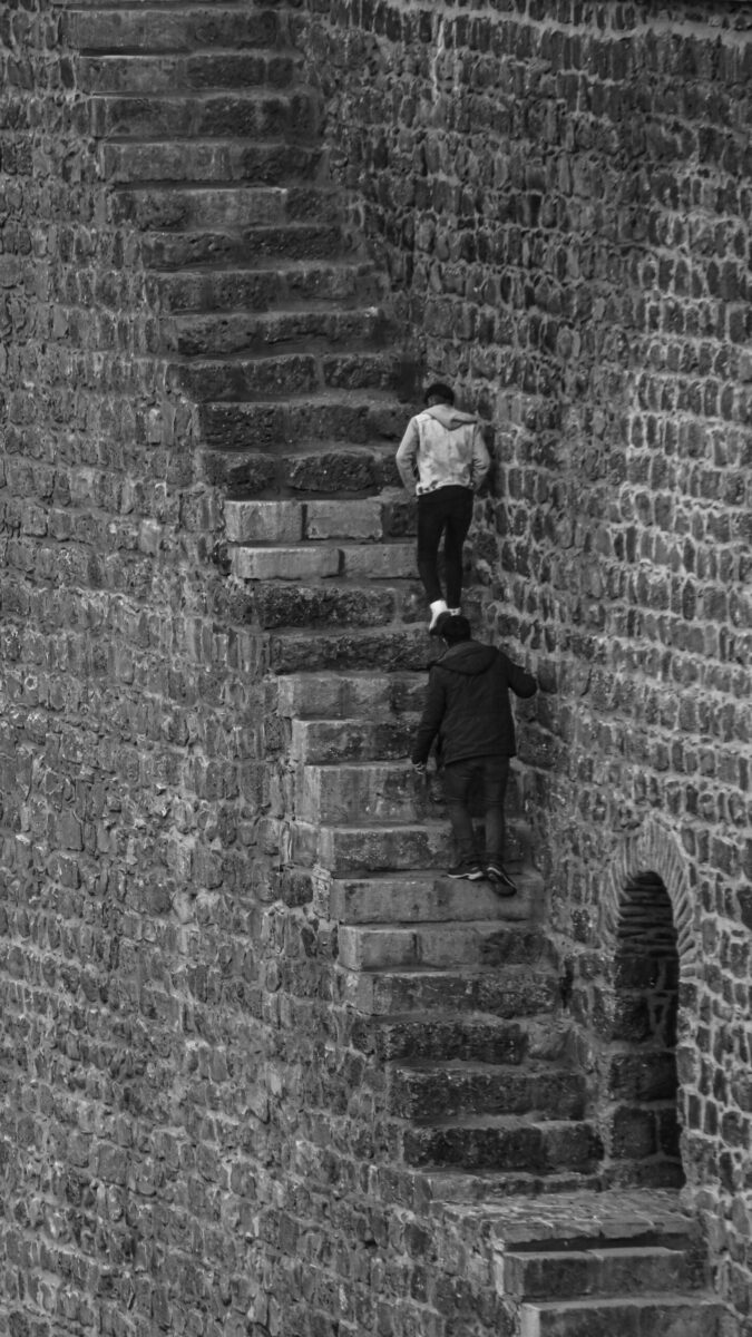 Two individuals climbing steep, narrow staircase on historical stone wall.